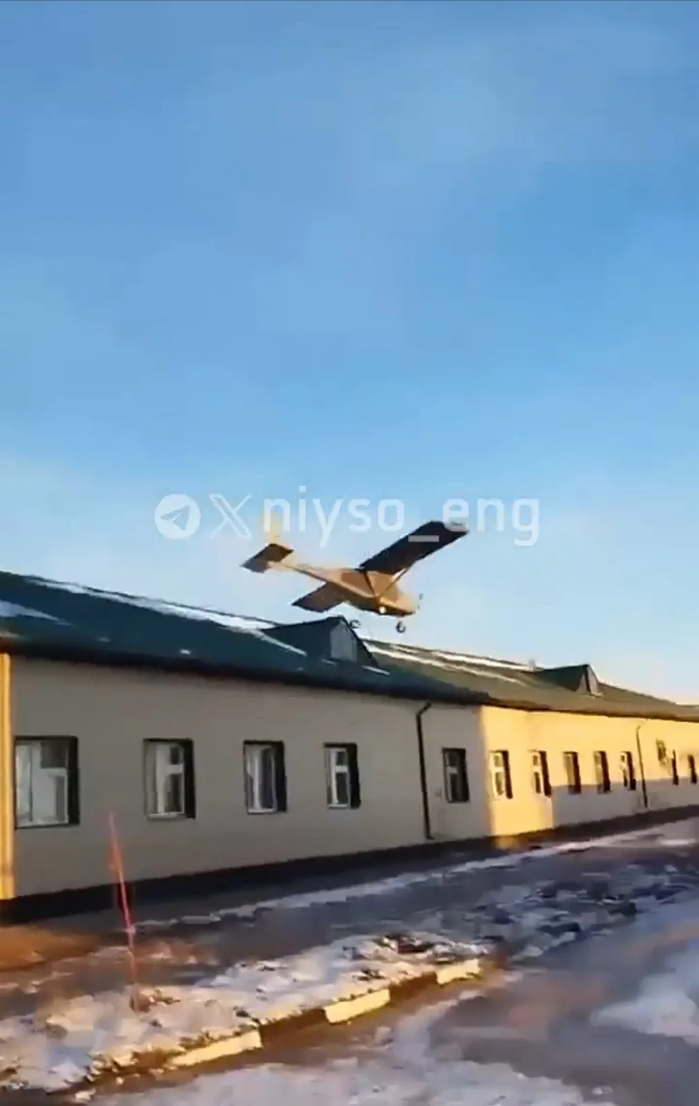 A small, single-engine airplane is captured mid-flight, flying extremely low just above the roof of a long, beige building…
