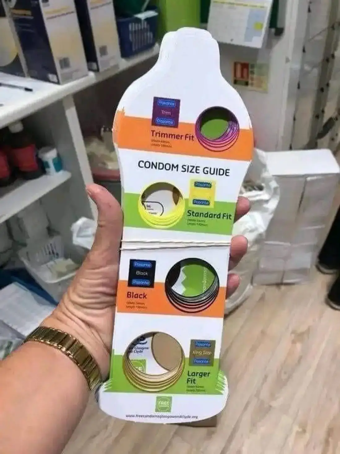 A hand holds a 'Condom Size Guide' display card showing four size options: Trimmer Fit, Standard Fit, Black, and Larger Fit…