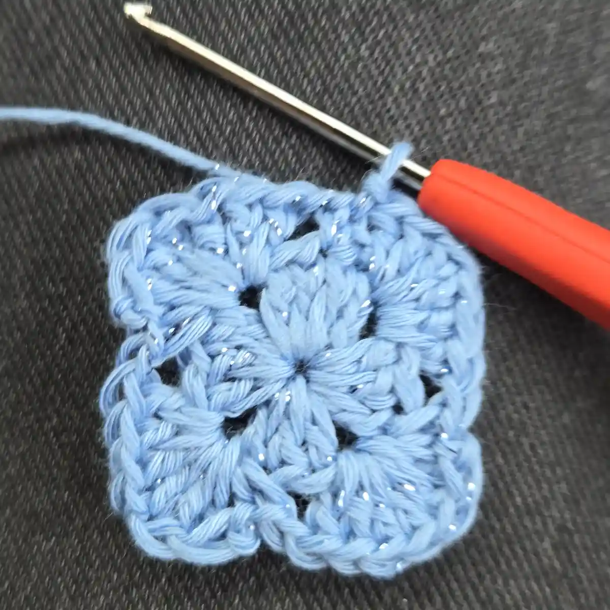 A close-up photo shows a small, light blue crocheted square with a textured, flower-like pattern, resting on a dark gray…