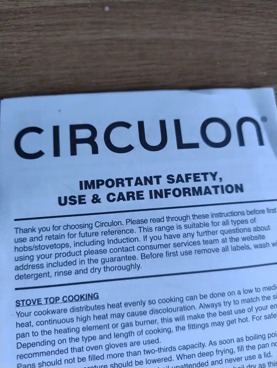 A close-up photo of a Circulon brand safety instruction sheet for cookware, resting on a wooden surface. The visible text…