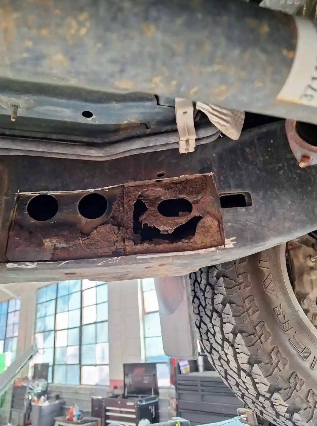 A close-up photo of the underside of a rusty vehicle shows a corroded metal panel with three holes on the left and a jagged…