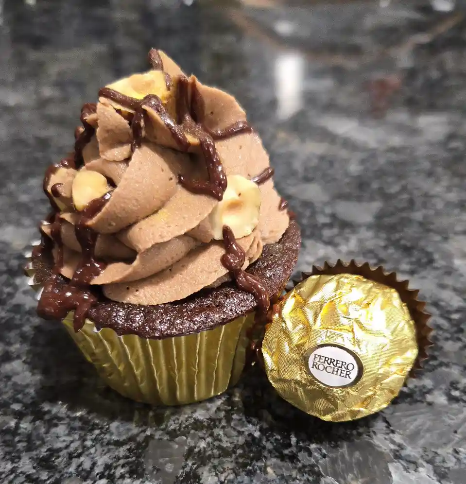 A chocolate cupcake with chocolate frosting, drizzled with chocolate sauce and topped with chopped hazelnuts sits next to an…