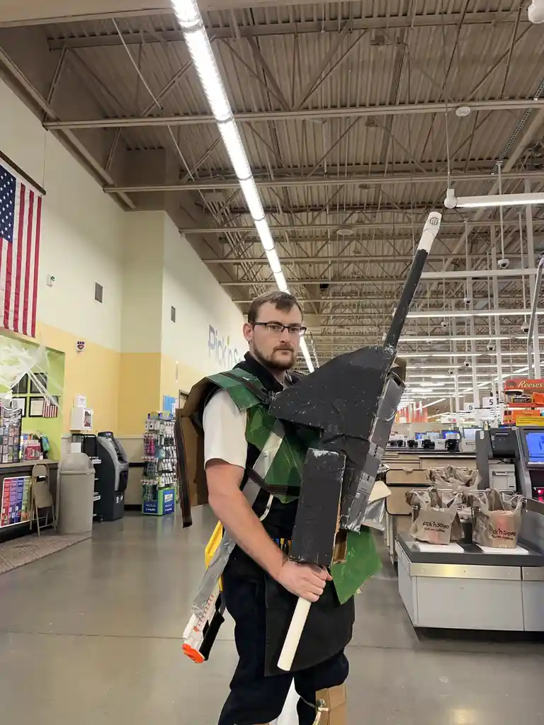 A man in a grocery store, wearing a homemade green and black cardboard costume resembling a Star Wars character, holds a…