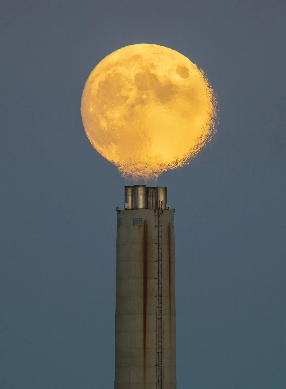 A large, golden full moon appears to be emerging from the top of an industrial smokestack, with its surface textured like a…