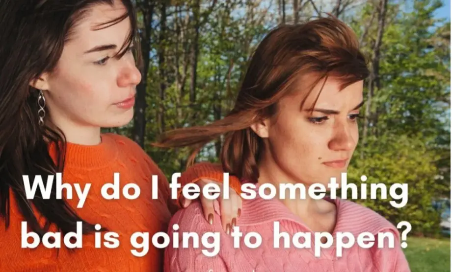 Two young women stand outdoors in a wooded area; one in an orange sweater has her arm around the other in a pink sweater…