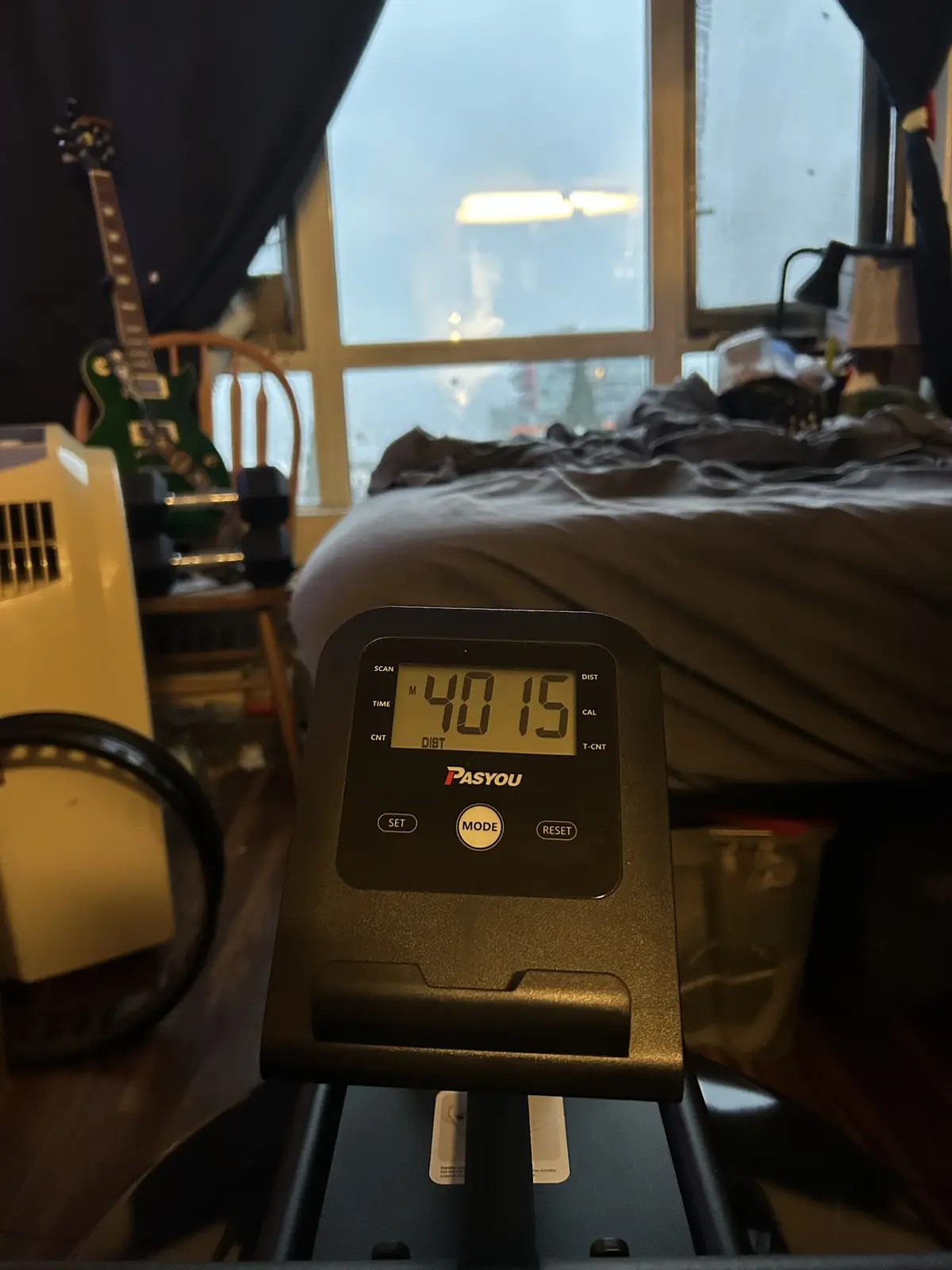 A close-up of a treadmill's digital display showing '40 15', with a messy bedroom background including a bed, a green…