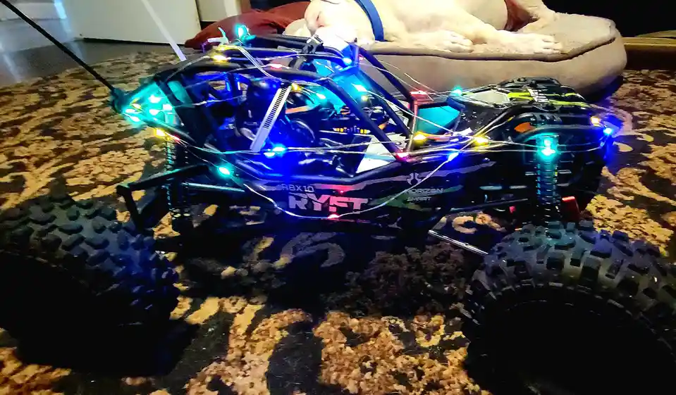 A black remote-controlled monster truck with glowing blue and green lights is parked on a patterned carpet. In the…