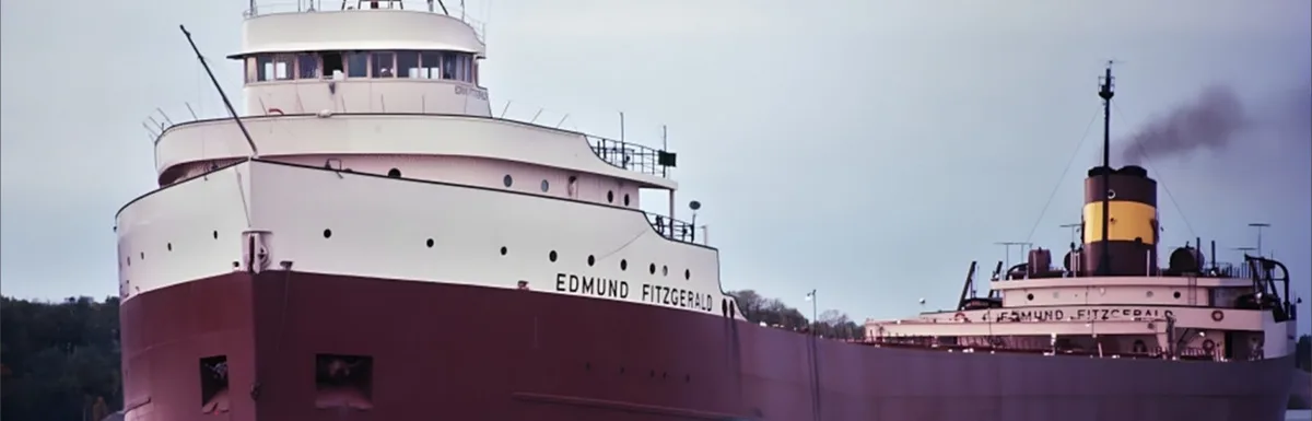 A wide shot of the freighter Edmund Fitzgerald, showing its massive hull painted white above and red below, with the name…
