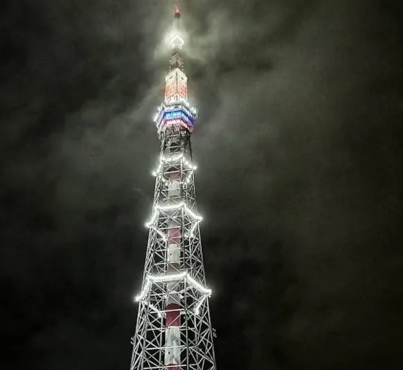 A tall, lattice-style tower, likely a communications or observation tower, is illuminated at night against a dark, cloudy…