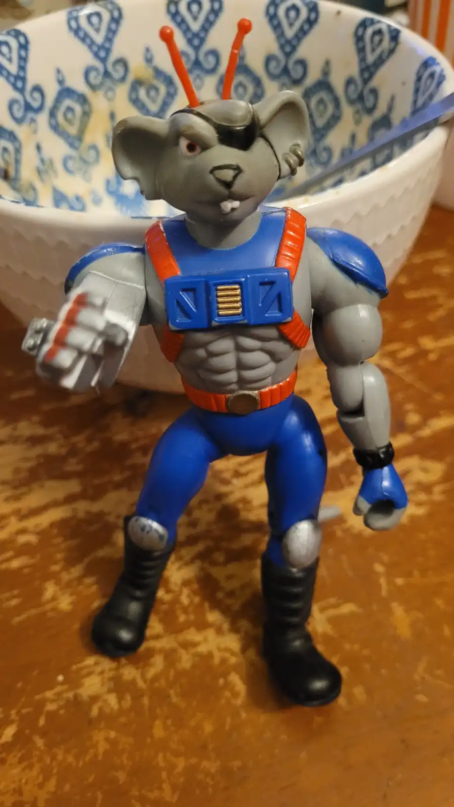 A gray plastic action figure of a muscular mouse with red antennae, an eyepatch, and blue/orange armor stands on a wooden…