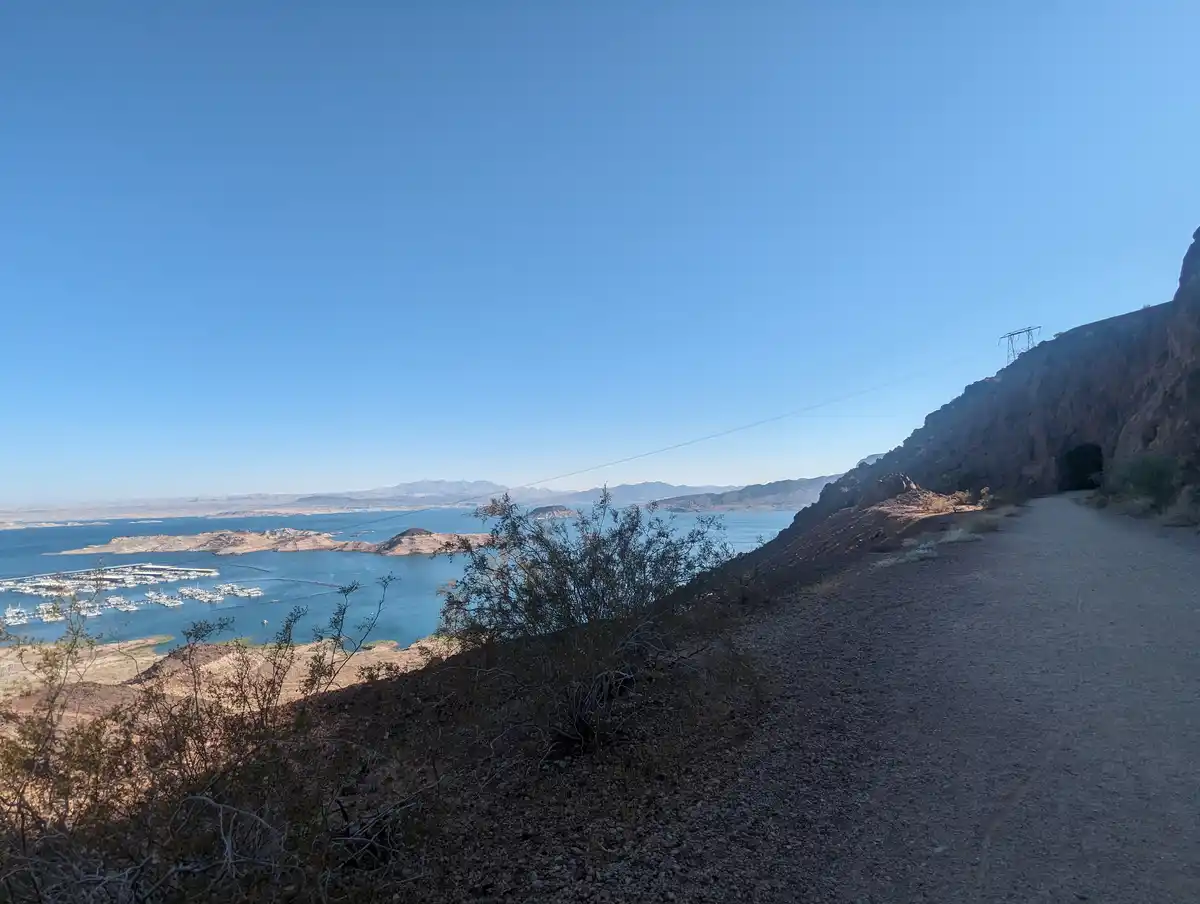 A scenic view of a desert landscape with a dirt path leading toward a tunnel in a rocky cliffside, overlooking a large blue…