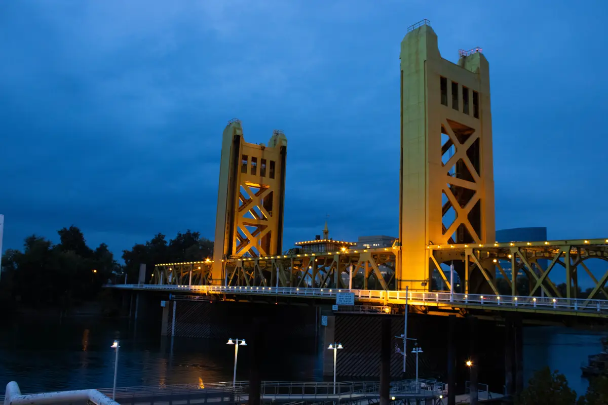 A photograph of the Tower Bridge in Sacramento, California, illuminated at dusk with warm yellow lights against a deep blue…
