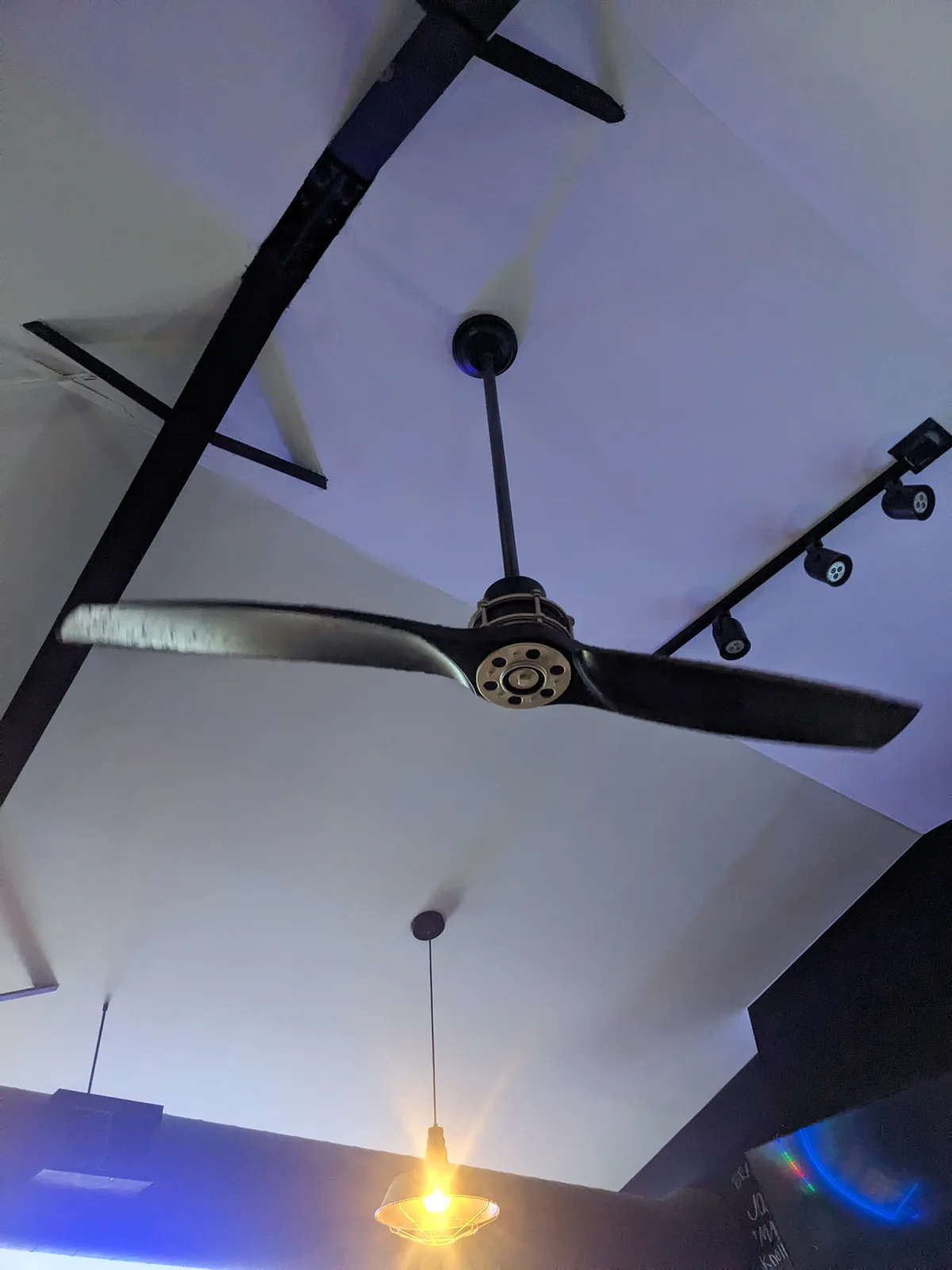 A low-angle photo looking up at a ceiling with a large black ceiling fan in motion, its blades blurred by speed. The fan is…