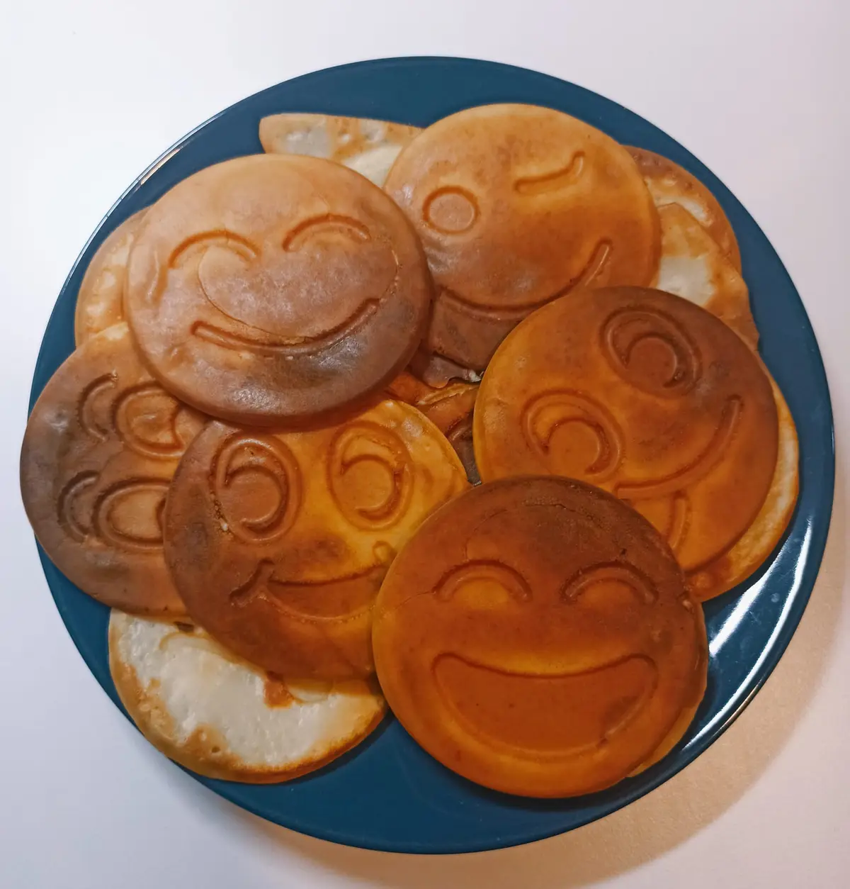 A blue plate holds a pile of golden-brown pancakes, each imprinted with different smiling emoji faces like winks, grins, and…