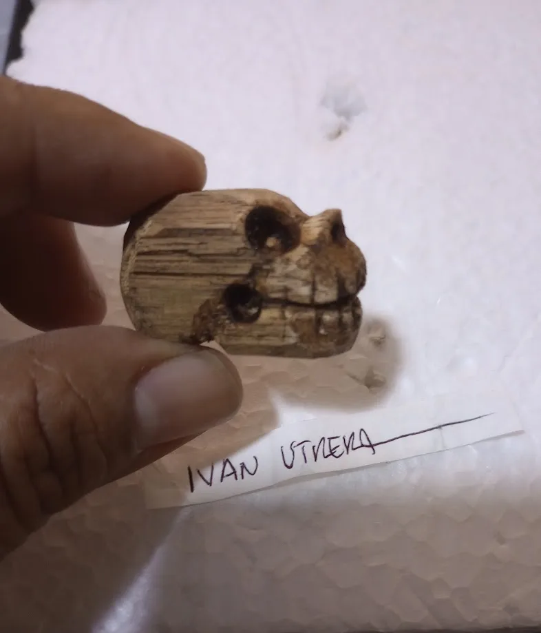 A hand holds a small, crudely carved wooden skull with dark, burnt-looking eyes and teeth. Below it, a piece of tape has…