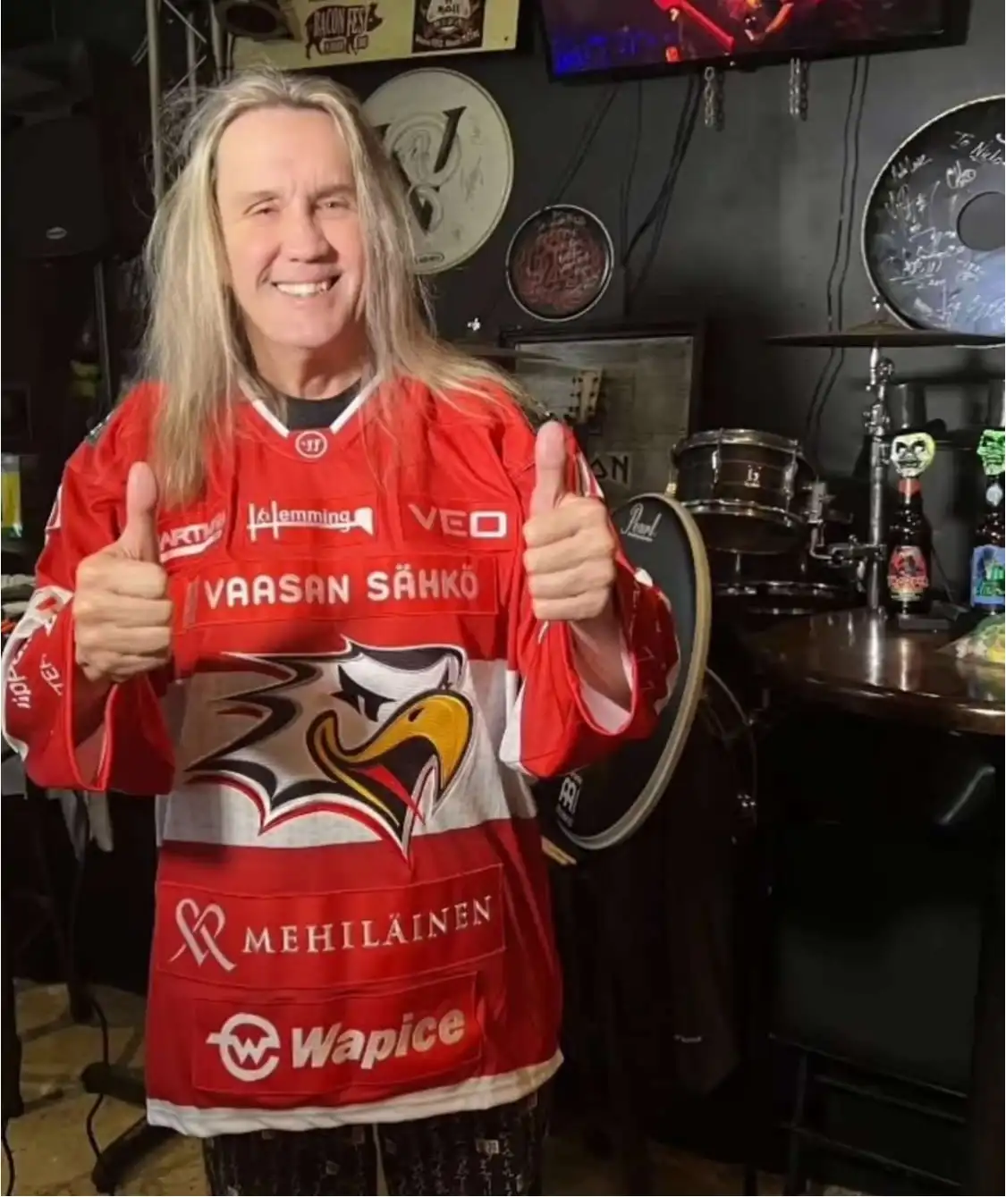 A man with long blonde hair wearing a red Finnish ice hockey jersey with 'VAASAN SÄHKÖ' across the chest, gives two thumbs…