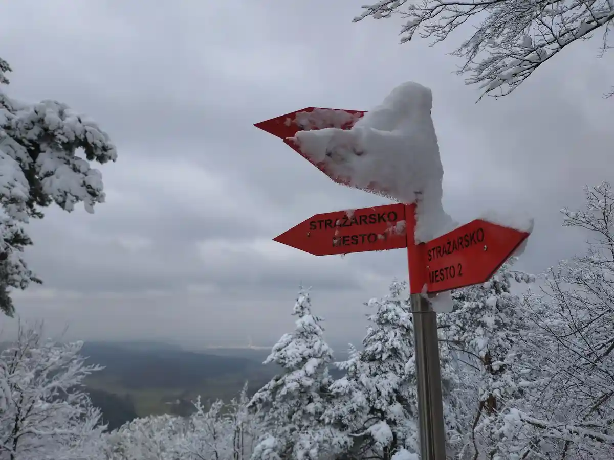 A snowy outdoor scene features a red directional signpost with two arrows pointing left and right, both labeled 'stražarsko…