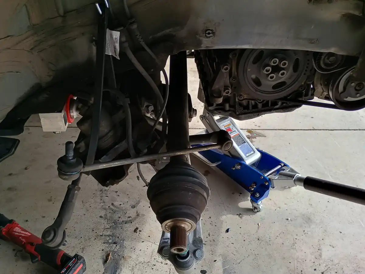 A low-angle photo shows the undercarriage of a car with a blue floor jack supporting it, exposing a newly replaced CV axle…