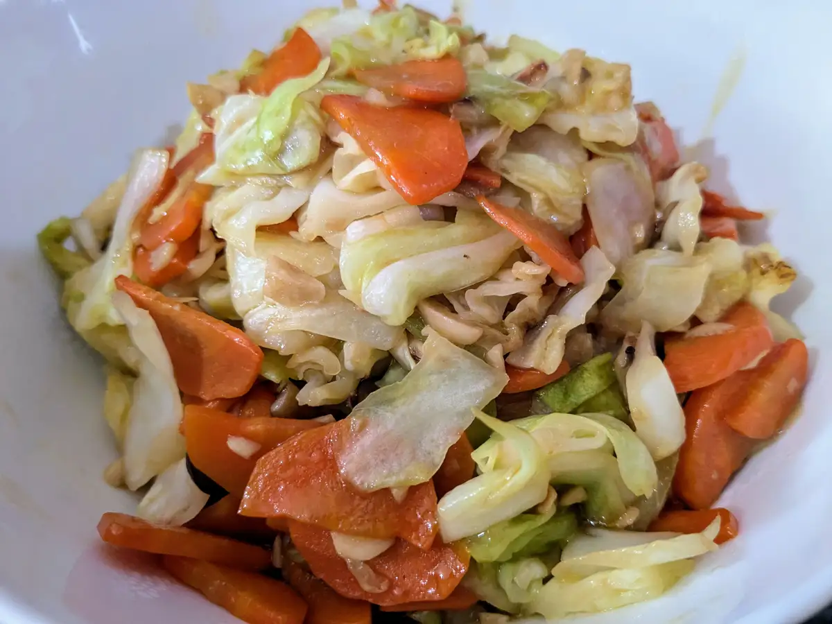 A close-up photo of a white bowl filled with stir-fried cabbage and carrot slices, glistening with sauce. The overlaid text…