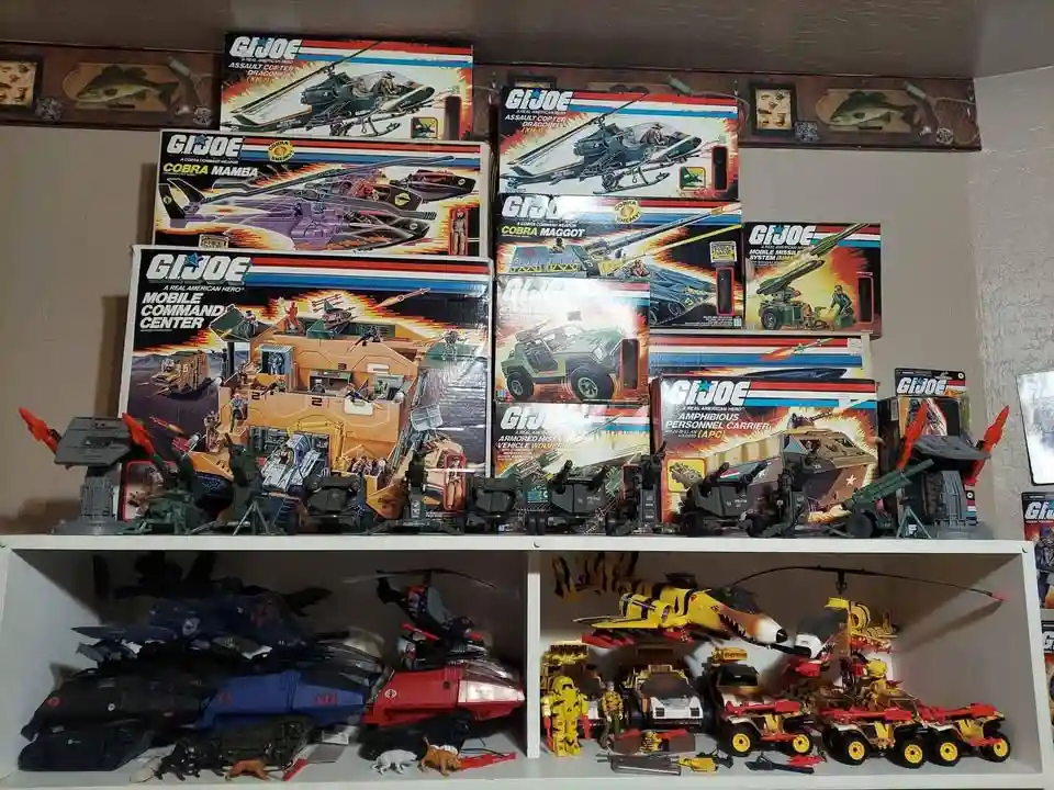 A shelf displays numerous vintage G.I. Joe toy boxes stacked on top, featuring vehicles like the Cobra Mamba and Mobile…