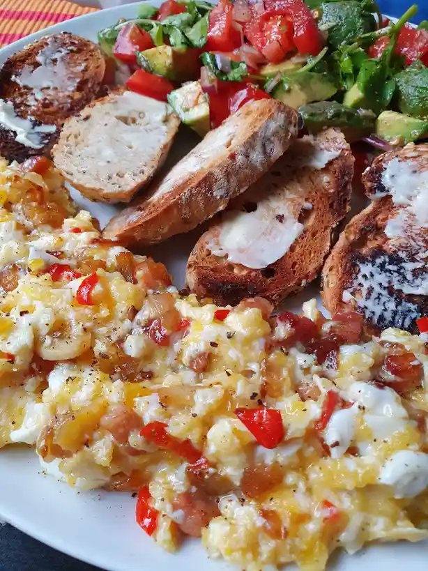 A close-up photo of a plate of breakfast food including scrambled eggs with red peppers and cheese, avocado and tomato…