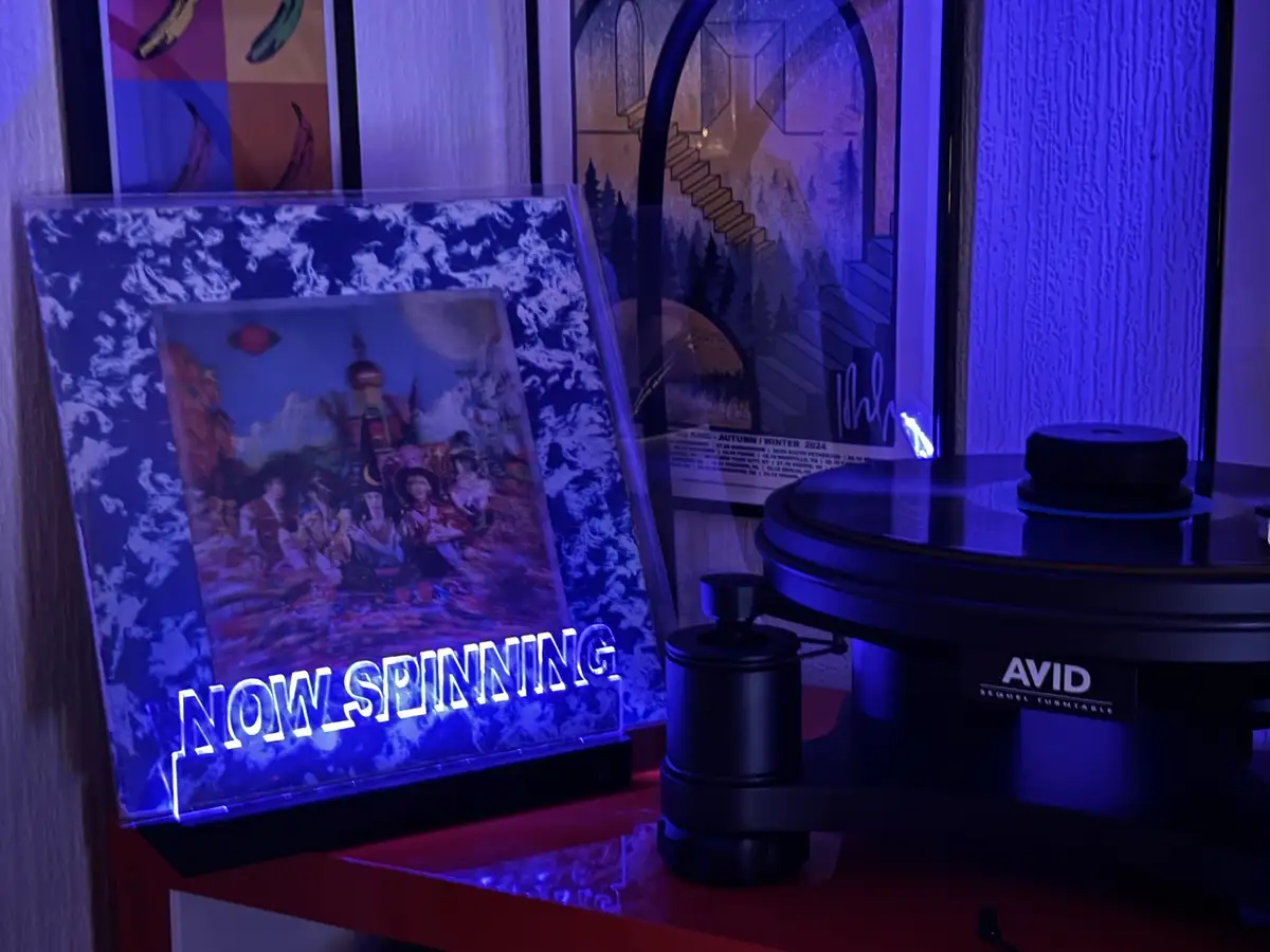 A vinyl record player with a glowing 'NOW SPINNING' sign sits on a red shelf next to a framed album cover under blue ambient…
