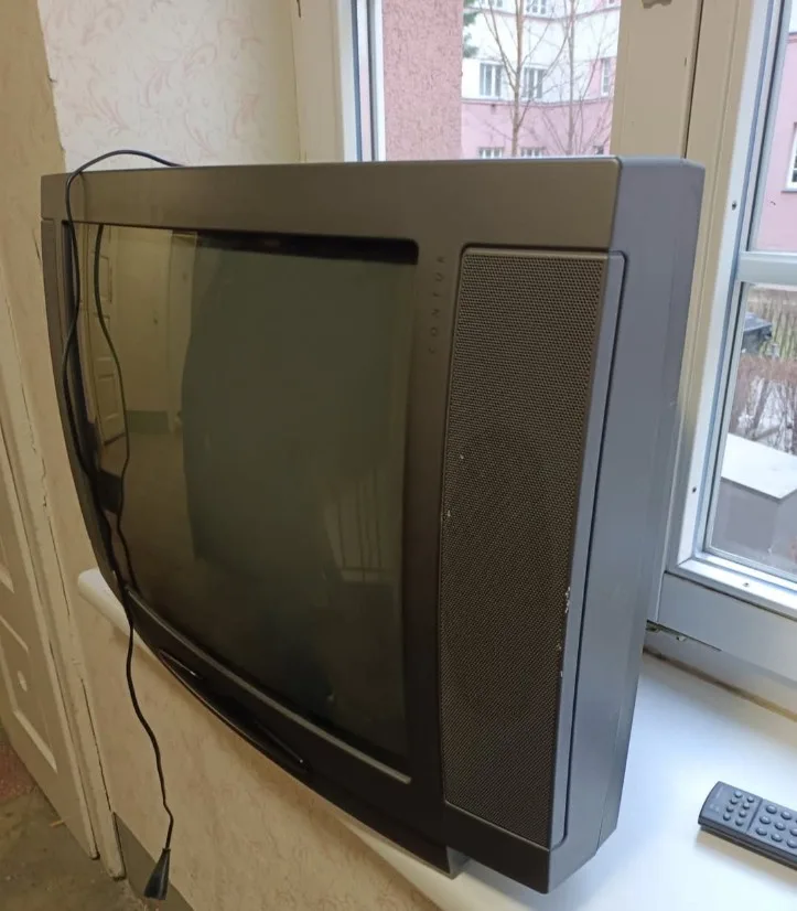 A black, boxy, old-style CRT television sits on a windowsill, its screen dark and reflecting the room. A black power cord…