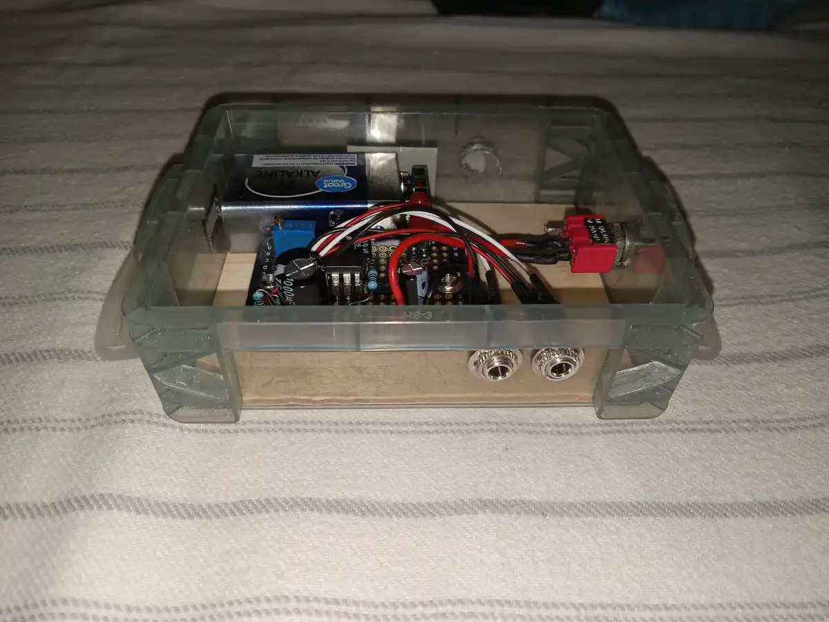 A homemade electronic device in a clear plastic case sits on a striped fabric surface, containing visible circuitry, wires…
