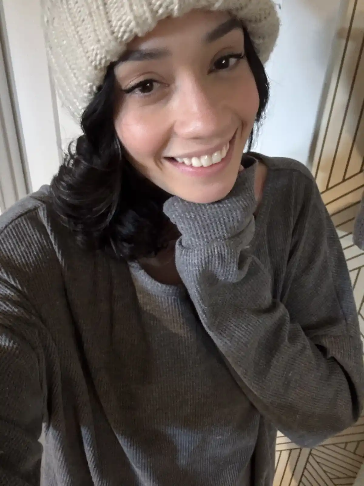 A woman with dark hair and a white knit beanie smiles warmly at the camera while wearing a grey turtleneck sweater. She has…