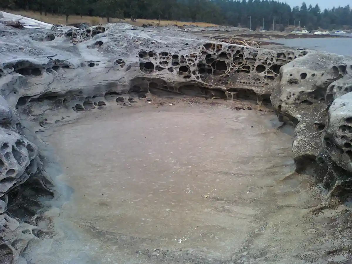 a natural rock formation with a shallow, bowl-shaped depression surrounded by porous, pitted rock walls, resembling a hot…