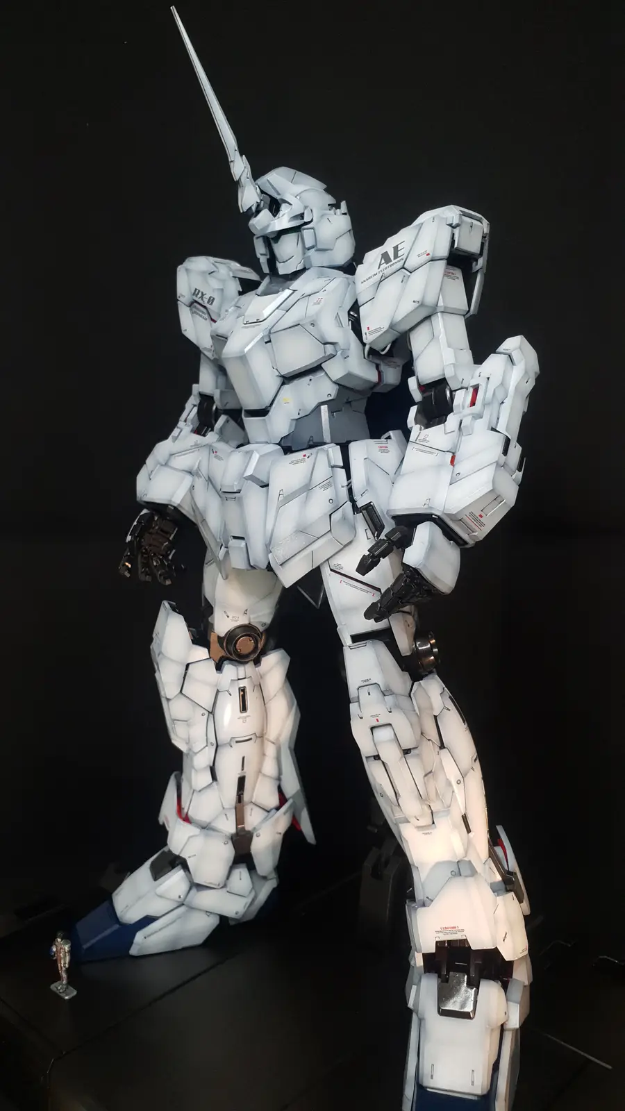 A large, detailed white and grey Gundam model, specifically the RX-0 Unicorn Gundam, stands against a black background with…