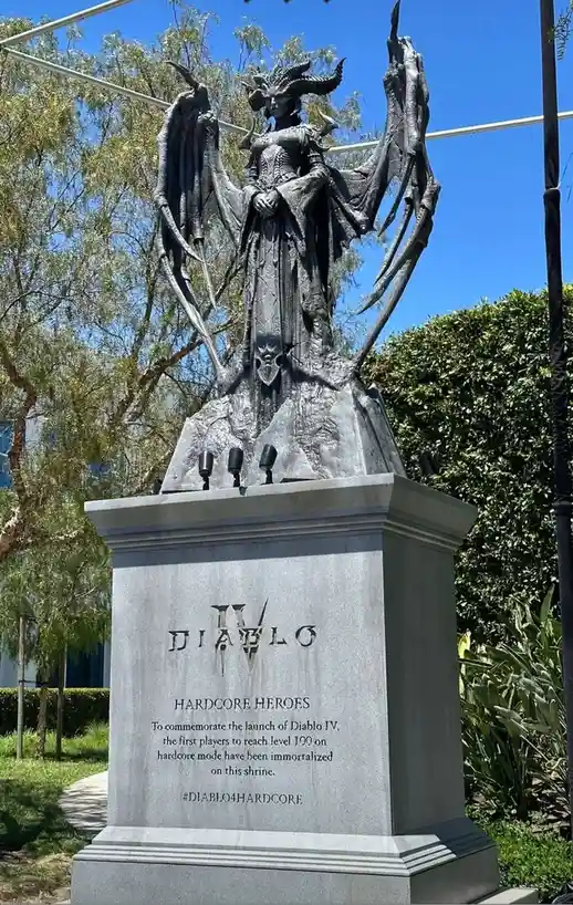 A large, dark statue of a winged demonic figure with horns stands atop a stone pedestal in a sunny outdoor setting. The…