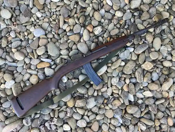 An AK-47-style rifle with a wooden stock and green sling lies diagonally across a bed of smooth, rounded river rocks. The…