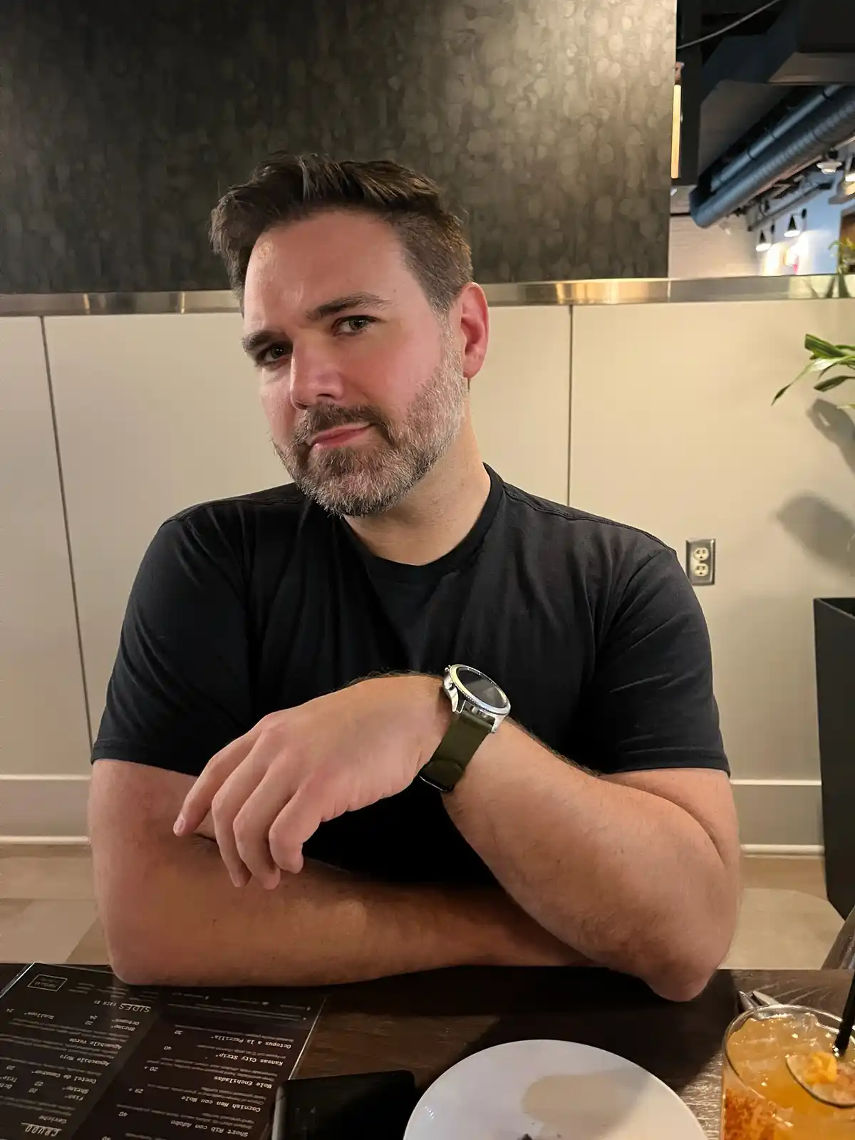 A man with a beard and black t-shirt sits at a restaurant table with arms crossed, looking directly at the camera with a…