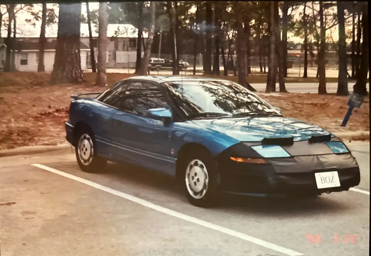 A blue 1990s-era Saturn SC coupe is parked in a lot with trees and a house in the background. The car has a custom black…