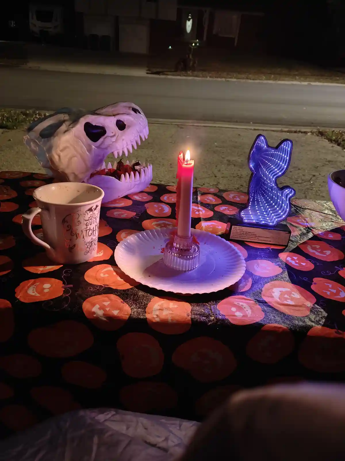 A nighttime outdoor table setup for Halloween, featuring a lit candle in a glass holder on a paper plate, a mug with 'Witch'…