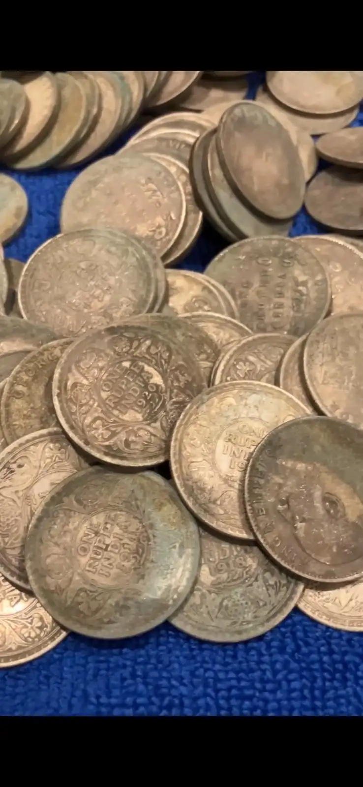 A pile of old, tarnished silver coins, mostly dated 1933 and marked 'ONE RUPEE INDIA', are scattered on a textured blue…
