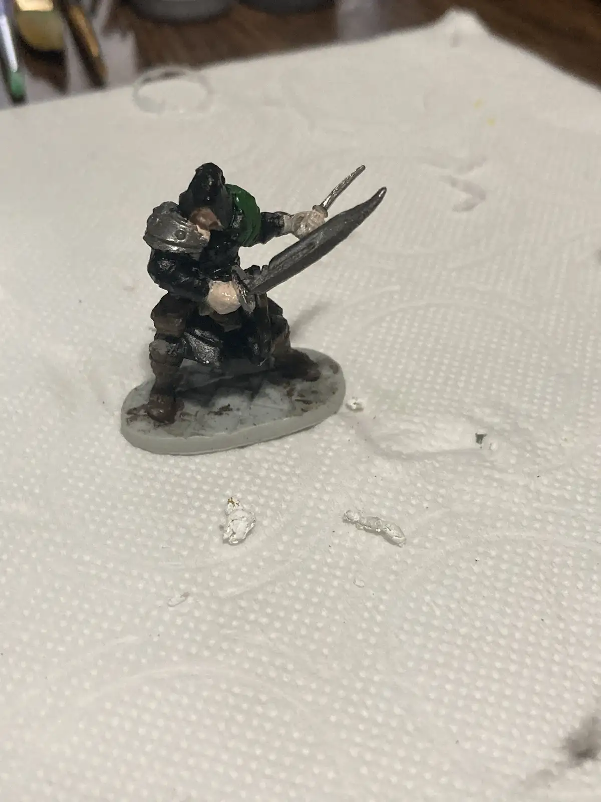 A painted miniature figurine of a warrior in dark armor with a green scarf, holding a sword and shield, stands on a white…