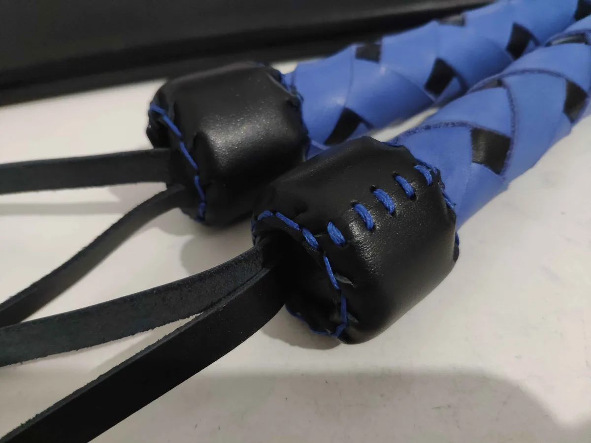 A close-up photo shows a handmade whip with a blue braided body and black leather handles, stitched with blue thread…