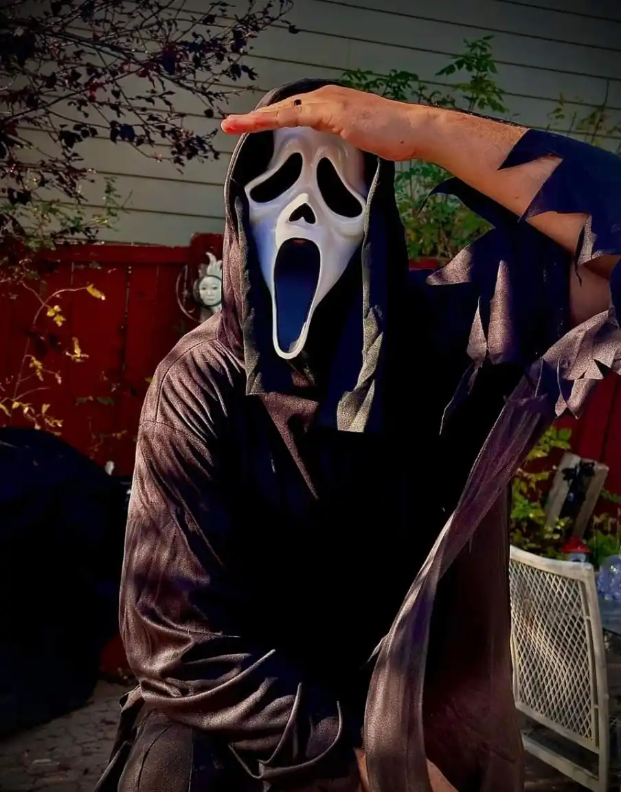 A person wearing a white Ghostface mask from the 'Scream' movie franchise and a dark, tattered hooded robe, stands outdoors…