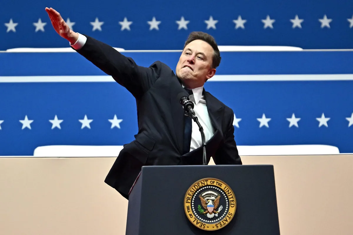 elon musk stands at a podium bearing the seal of the president of the united states, gesturing with his right arm raised…