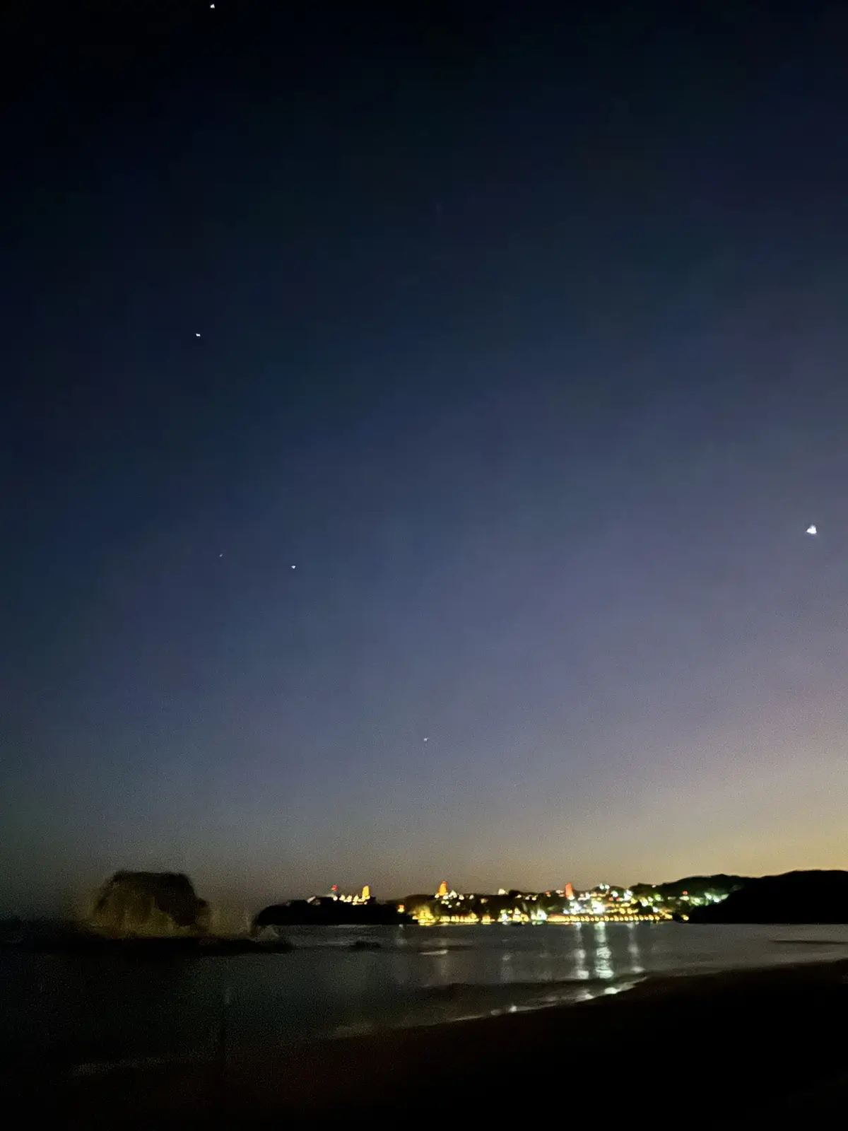 A night photograph of a coastal scene with a dark sky, faint stars, and a distant illuminated shoreline reflected in calm…