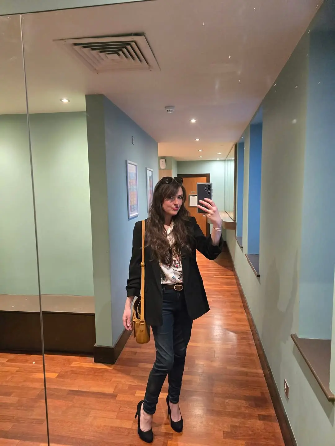 A woman with long brown hair and sunglasses perched on her head takes a mirror selfie in a hallway with light blue walls and…