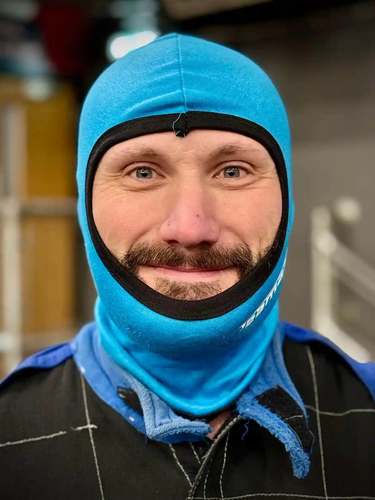 A man with a mustache and green eyes wears a bright blue balaclava that covers his head and neck, leaving only his eyes…