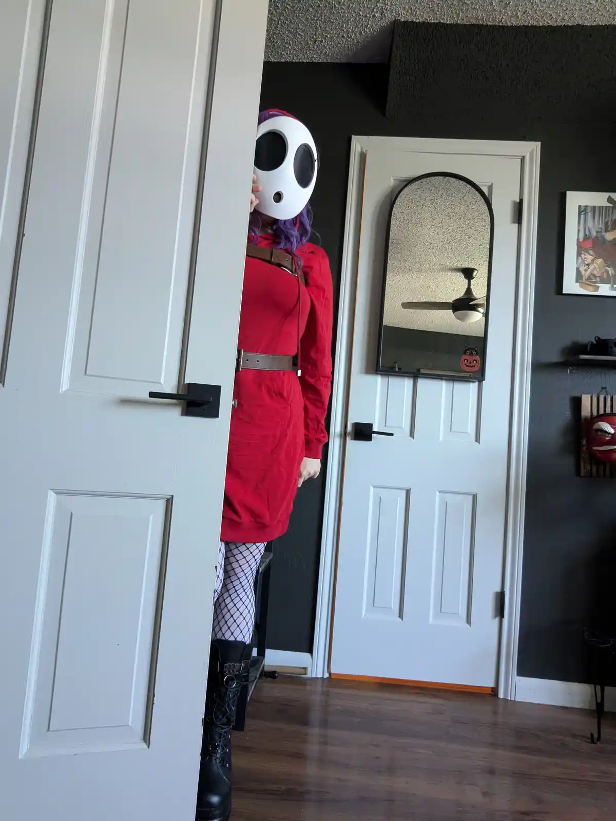 A person in a red dress, fishnet stockings, and black boots peeks from behind a white door, wearing a white mask with large…