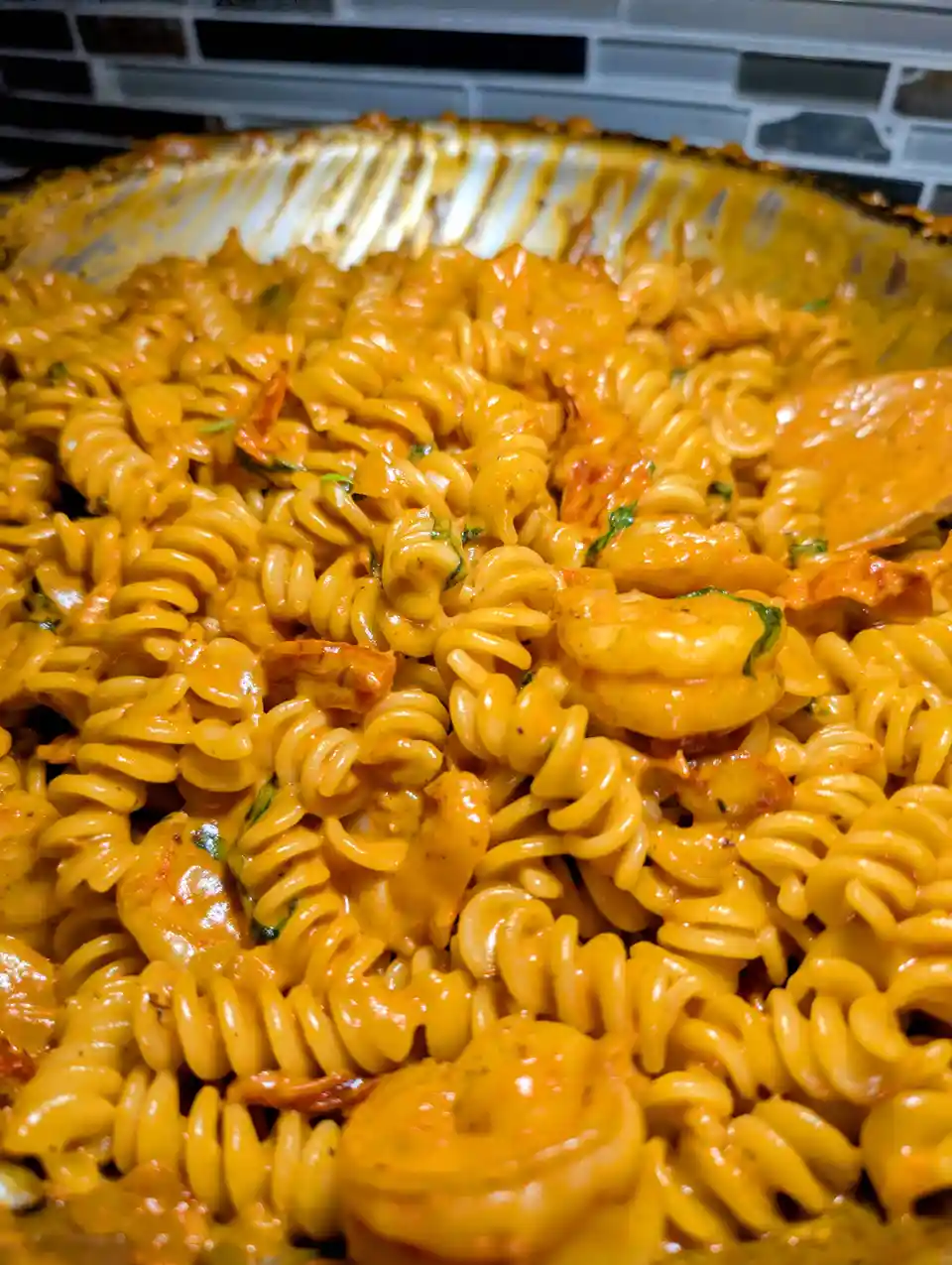 A close-up photo of a baking dish filled with rotini pasta coated in a thick, orange-red sauce, mixed with chunks of chicken…