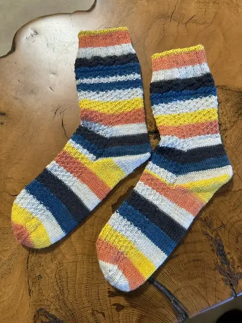 A pair of hand-knitted socks with vibrant horizontal stripes in blue, yellow, orange, and white are laid out on a rustic…