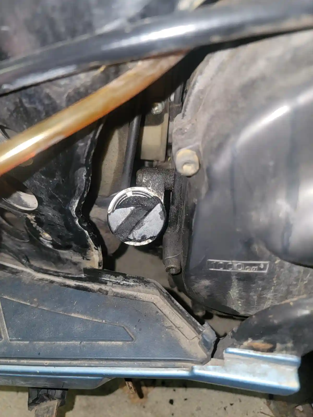A close-up photo of a motorcycle's engine area, showing a dirty oil cap with a visible '1000 cm³' engraving on the engine…