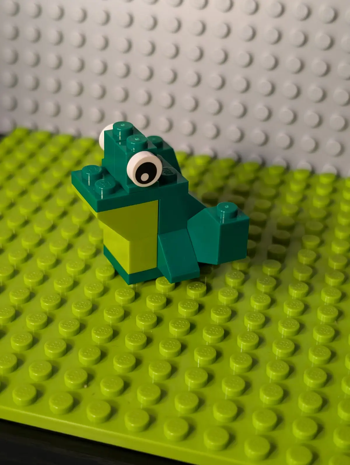 A small, crudely built frog made of green and teal Lego bricks sits on a lime-green Lego baseplate, with a single large…