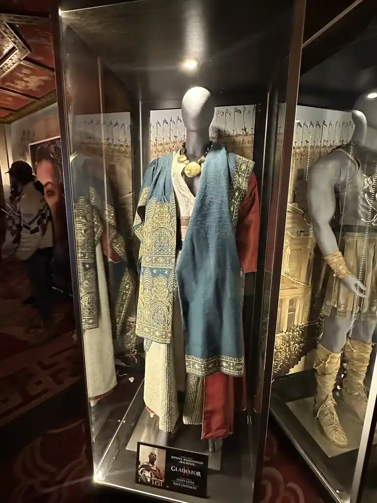 A museum display case shows a mannequin dressed in ornate, layered Roman-style clothing including a blue patterned robe…